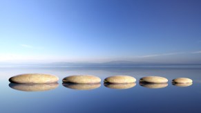 Zen stones row from large to small  in water with blue sky and peaceful landscape background.