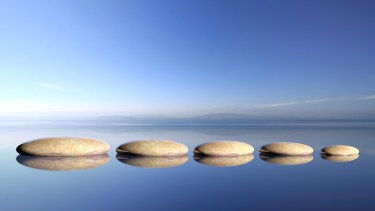 Zen stones row from large to small in water with blue sky and peaceful landscape background.