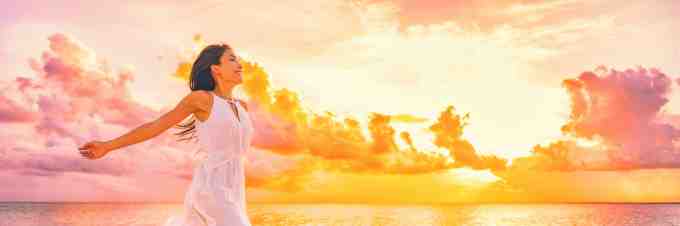 Well being free woman with open arms in the air blissful happiness concept banner. Happy woman against pink pastel colorful sunset sky.